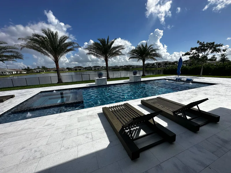 Pool and spa on a lake lot, with black chaise loungers and palms along the fence