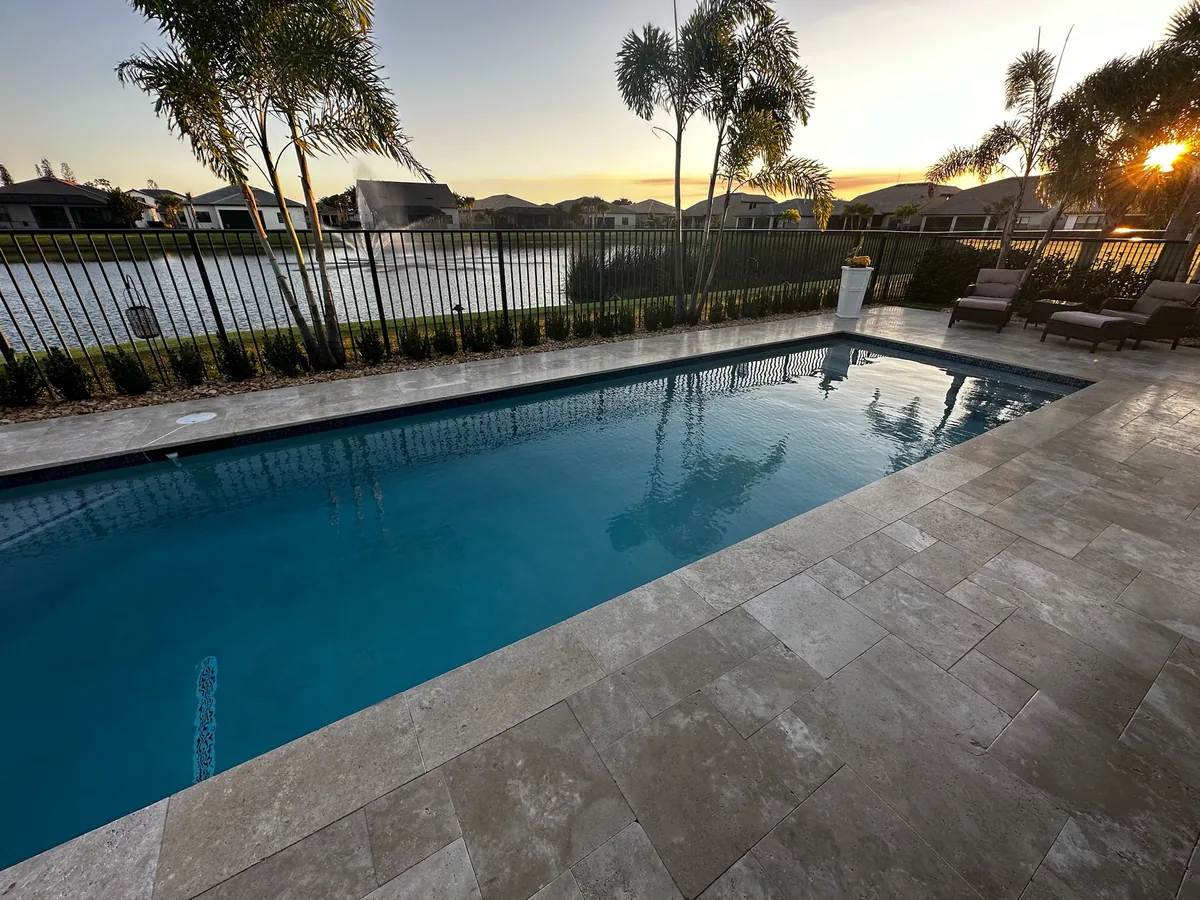 Pool with a light stone deck at sunset, a fence and a lake behind it