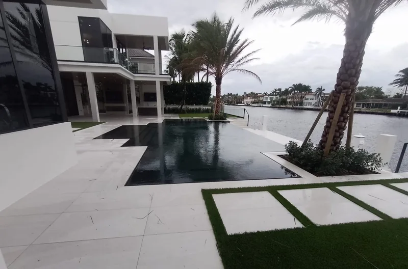 Dark-finished pool with a low edge, stepping pavers and palms beside a waterway