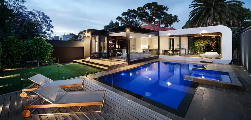 Lit pool and spa at night beside a modern pavilion, with loungers on a wood deck