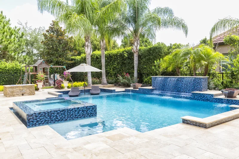 Custom pool with a raised glass-tile spa spilling into it, a sun shelf and travertine deck, framed by palms