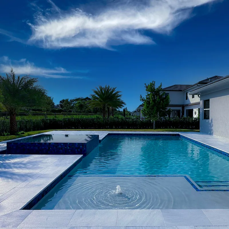 Pool with a bubbler on the shelf, palms and a white home under a blue sky