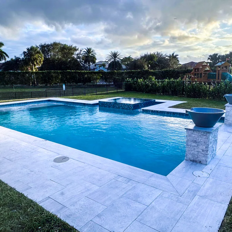 Pool with a fire bowl on a stone pillar and a white travertine deck at dusk