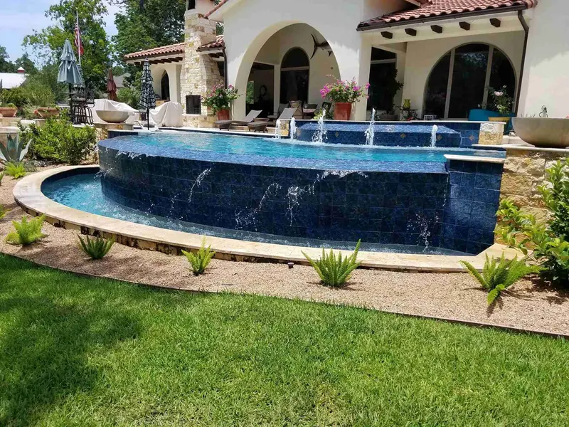 Raised spa with a vanishing edge spilling down a glass-tile wall in front of a Mediterranean-style home