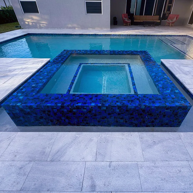 Raised square spa faced in dark blue glass mosaic tile beside a pool with light stone decking